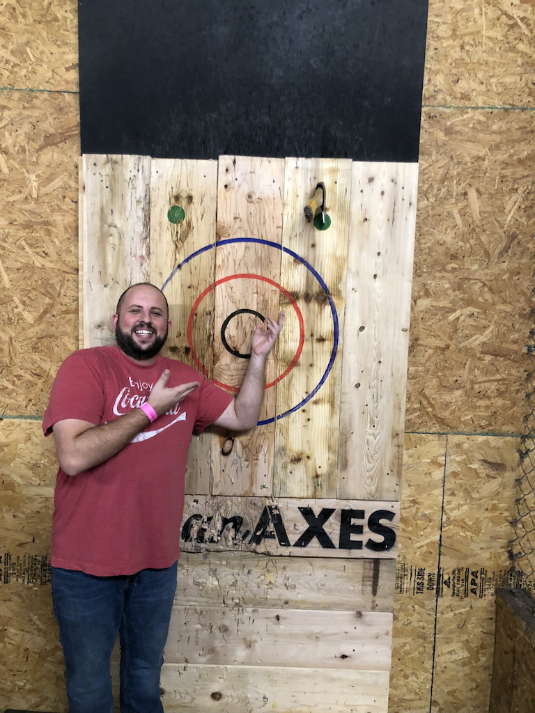 A man in a victory stance, standing in front of a target for axe throwing with the axe sticking in the "Clutch" circle