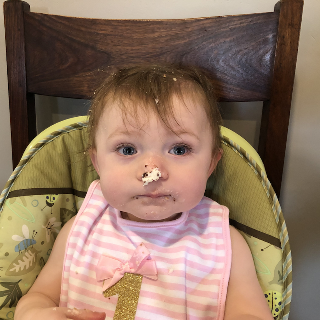 A young baby girl sitting in a high chair covered in what looks like birthday cake and frosting